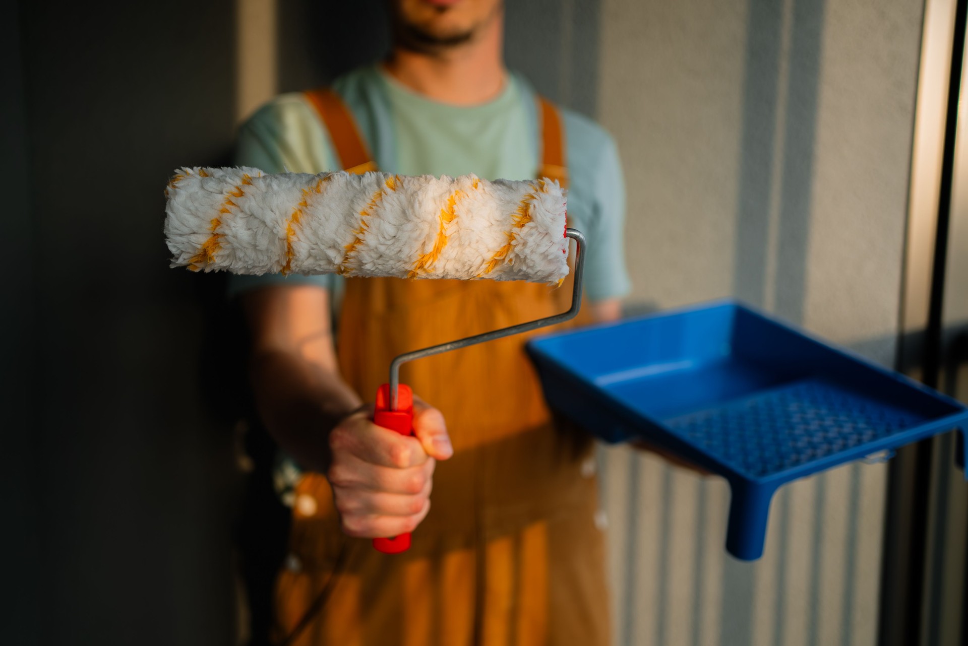 Professional painter gripping paint roller near blue tray, preparing interior wall renovation with natural light casting dramatic shadows across workspace